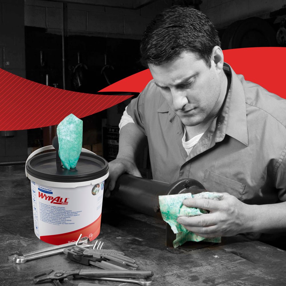 A man in work clothes cleans a metal pipe with a green WypAll® wipe from Kimberly-Clark GmbH. A carton (6 dispenser buckets) of wipes is placed among tools. The photo is mostly black and white, except for the wipes and the label.