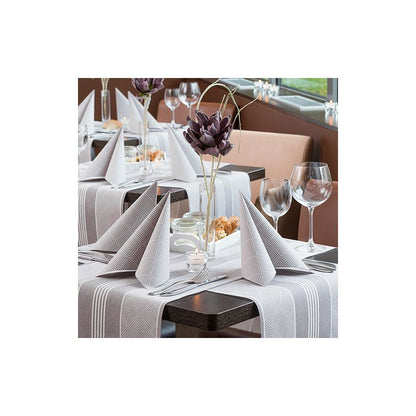 A beautifully set dining table with folded napkins and wine glasses on a striped tablecloth from the ROYAL collection of PAPSTAR GmbH. The PAPSTAR fabric-like table runner PV-tissue mix adds elegance to the scene, while decorative flowers and bread baskets complete the arrangement. Chairs and windows can be seen in the background.