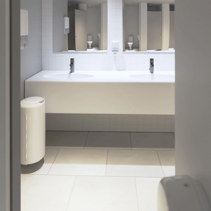 A modern public restroom features two white sinks with rectangular mirrors above, surrounded by smooth light gray tiles. A Tork 561000 Mini Dispenser for Liquid Soap Elevation S2 from Essity Professional Hygiene Germany GmbH is mounted next to the sinks, along with a white trash bin to ensure cleanliness.