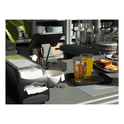 In a counter scene in a café or kitchen, a Tork Xpressnap Fit® 272901 countertop napkin dispenser from TORK - Essity Professional Hygiene Germany GmbH is presented next to the cash register. Next to it, on a tray, are two glasses of orange juice and a stack of cookies, while bottles and dishes are displayed on shelves in the background.
