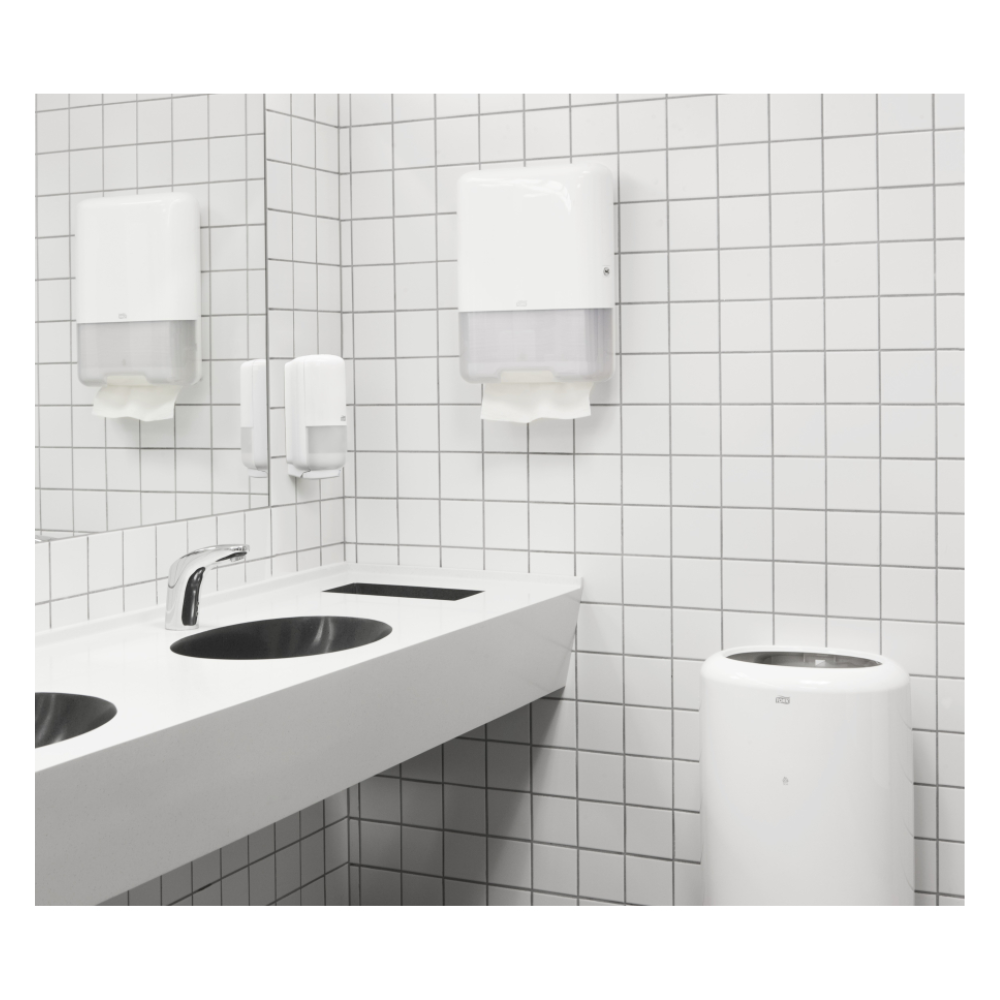 Eine moderne öffentliche Toilette verfügt über ein weißes Waschbecken mit zwei Wasserhähnen, eine weiß geflieste Wand und einen TORK Tork 100278 extraweiche Zickzack-Handtücher Premium H3 2-lagig | Karton (15 Packungen) Spender mit hochwertigen 2-lagigen Papierhandtüchern. Ergänzt durch einen Seifenspender und einen Mülleimer ist die Umgebung sauber und minimalistisch.