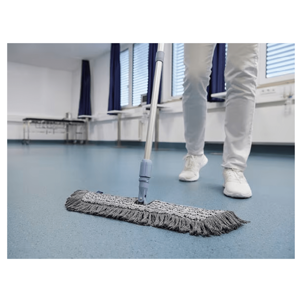 A person in white shoes and pants is mopping a blue floor with a Vileda GmbH Professional Swep Duo r-MicroCombi Mop - 50 cm in a room with closed blinds and blue curtains. A folding table stands nearby and a radiator is under the windows.