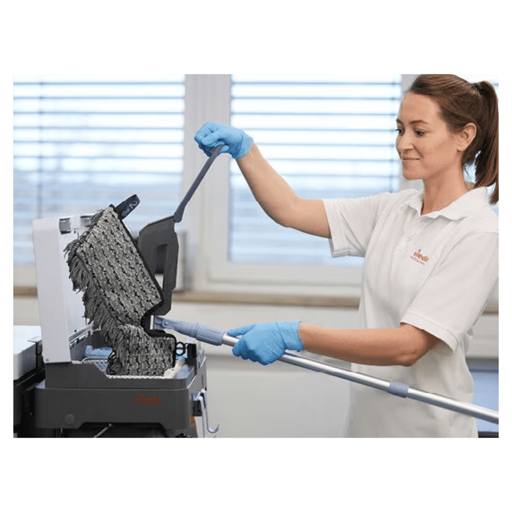 A person with blue gloves and a white shirt is cleaning a textured black surface with the Vileda Professional Swep Duo r-MicroCombi Mop - 50 cm from Vileda GmbH. The scene appears to be indoors, with a large window in the background through which natural light enters the room.
