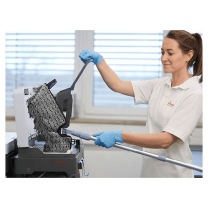 A person with blue gloves and a white shirt is cleaning a textured black surface with the Vileda Professional Swep Duo r-MicroCombi Mop - 50 cm from Vileda GmbH. The scene appears to be indoors, with a large window in the background through which natural light enters the room.