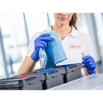 A person with a white shirt and blue gloves holds a blue Vileda Professional r-MicroTuff Swift microfiber cloth from Vileda GmbH over a row of containers with differently colored lids; the scene illustrates the efficient cleaning task using the four-color system for organization.