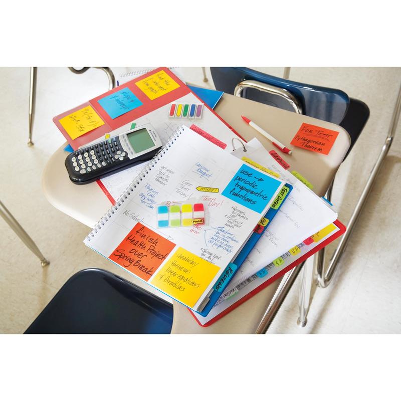 On a classroom desk lie notebooks, a planner with 3M Post-it® Index Strong Filing Tabs (Mini, yellow, red, blue & green), calculator, pens, highlighters & folders; empty chairs surround it and the tabs help organize documents.