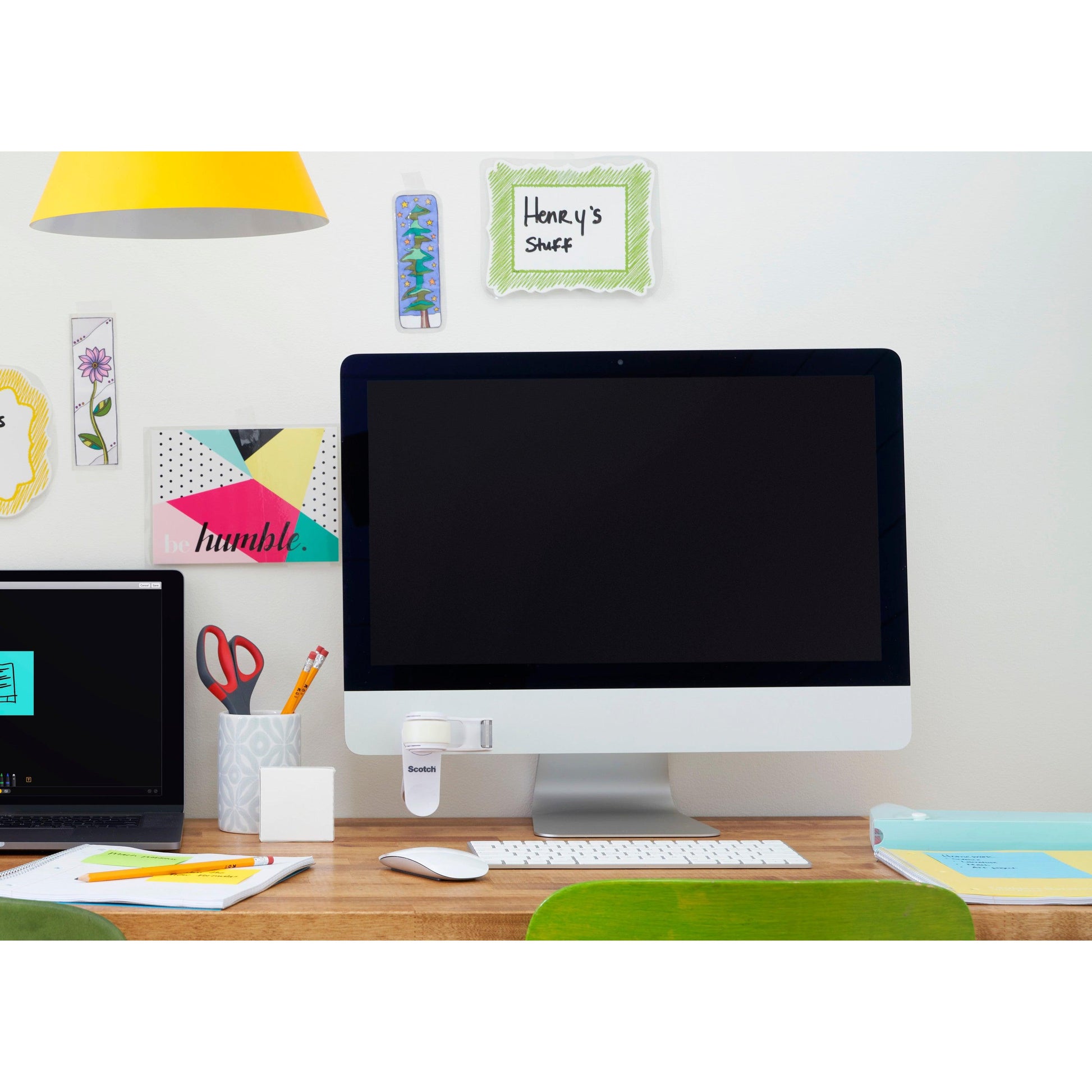 Ein aufgeräumter Schreibtisch mit einem großen Monitor, einem geschlossenen Laptop, einem Becher mit Stiften und Scheren, Büromaterial und dem 3M Scotch™ Clip & Twist Klebeband-Abroller C19 mit Magic™ Unsichtbarem Klebeband auf dem Display. An der Wand befinden sich dekorative Notizen wie "Henry's Stuff".